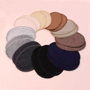 Op de afbeelding: Een verzameling ovale schoudervullingen in verschillende kleuren, waaronder beige, grijs, bruin, zwart en marineblauw. De vullingen zijn in een cirkelvormig patroon gerangschikt tegen een zachtroze achtergrond. Deze vullingen zijn waarschijnlijk voor kleding.