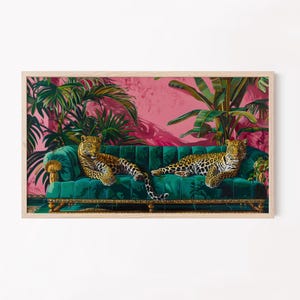 May include: A framed art print featuring two leopards resting on a teal green velvet sofa. The background includes pink walls and lush tropical plants. The artwork is enclosed in a light wood frame.
