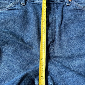May include: Close-up of blue denim jeans with a yellow measuring tape showing a length of approximately 13 inches. The jeans have classic five-pocket styling with copper rivets and a button closure.