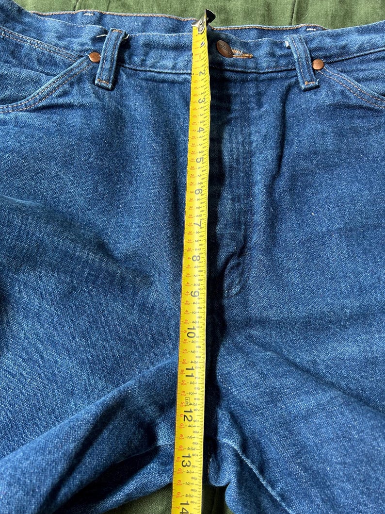 May include: Close-up of blue denim jeans with a yellow measuring tape showing a length of approximately 13 inches. The jeans have classic five-pocket styling with copper rivets and a button closure.