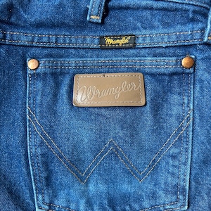 May include: Close-up of blue denim jeans featuring a back pocket with the brand name "Wrangler" on a leather patch. The pocket has a distinctive "W" stitching design. The jeans have copper rivets and a small label with the brand name.
