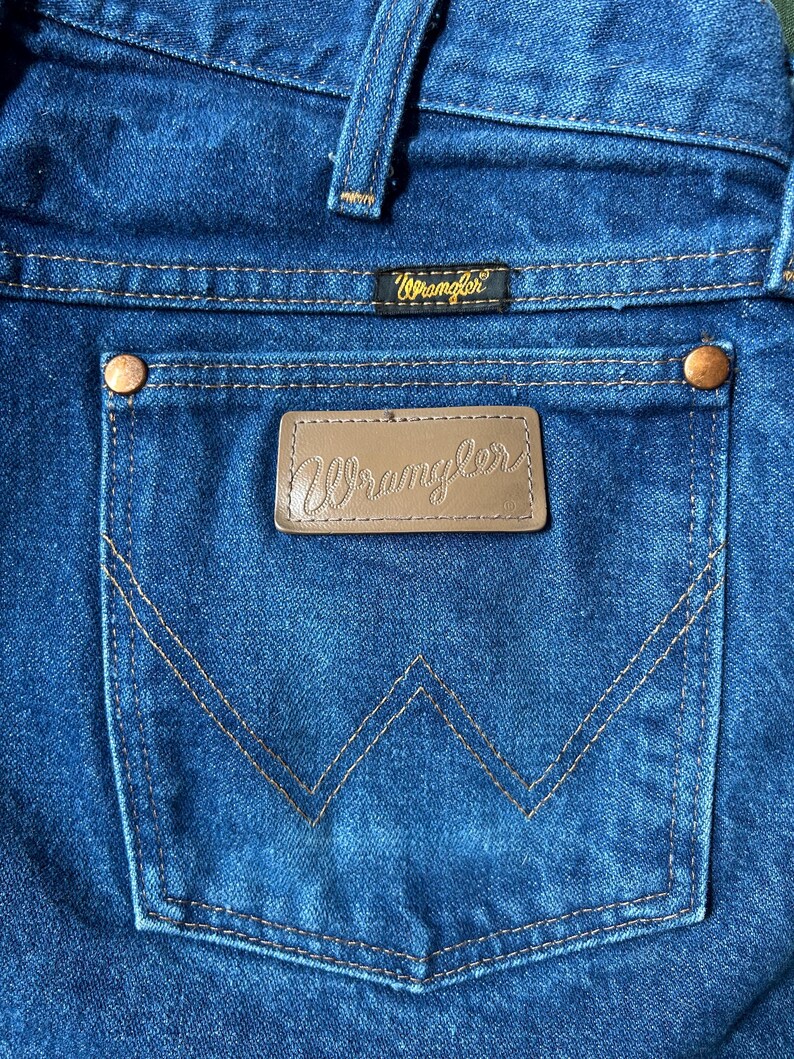 May include: Close-up of blue denim jeans featuring a back pocket with the brand name "Wrangler" on a leather patch. The pocket has a distinctive "W" stitching design. The jeans have copper rivets and a small label with the brand name.