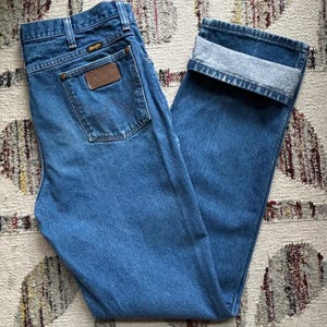 Pu&ograve; includere: Jeans blu in denim con gamba dritta e risvolto. I jeans presentano una toppa in pelle marrone sulla tasca posteriore con il marchio "Wrangler". I jeans sono realizzati in denim di media tonalit&agrave;.