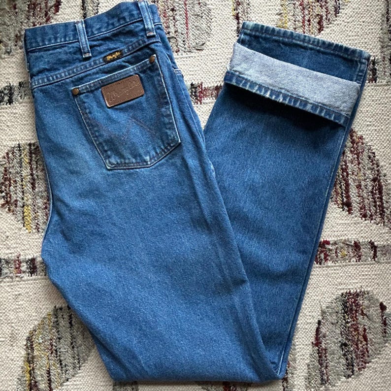 Pu&ograve; includere: Jeans blu in denim con gamba dritta e risvolto. I jeans presentano una toppa in pelle marrone sulla tasca posteriore con il marchio "Wrangler". I jeans sono realizzati in denim di media tonalit&agrave;.