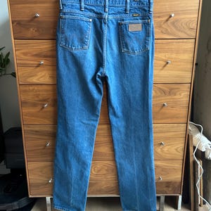 Pu&ograve; includere: Jeans blu in denim con una classica vestibilit&agrave; a gamba dritta. I jeans presentano due tasche posteriori e una piccola toppa in pelle marrone con il nome del marchio. I jeans sono appesi a una gruccia di legno.