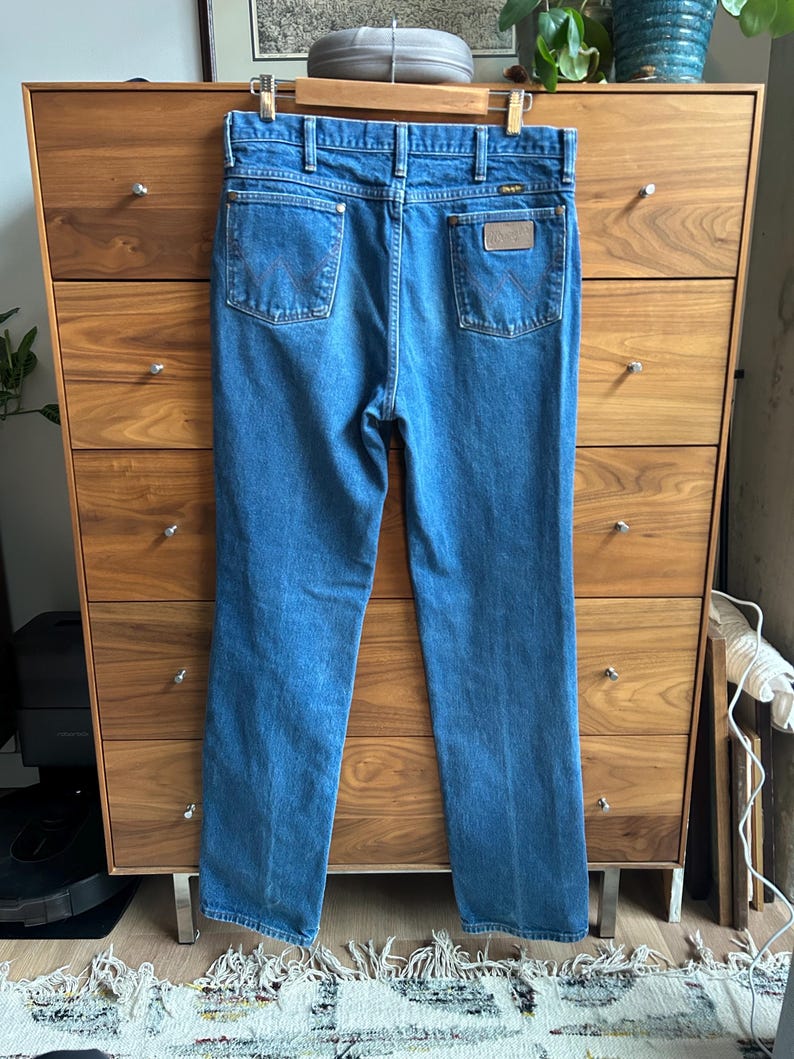 Pu&ograve; includere: Jeans blu in denim con una classica vestibilit&agrave; a gamba dritta. I jeans presentano due tasche posteriori e una piccola toppa in pelle marrone con il nome del marchio. I jeans sono appesi a una gruccia di legno.