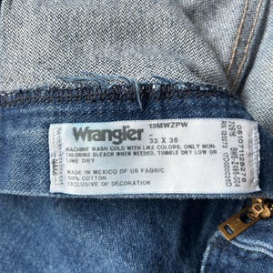 May include: Close-up of a pair of blue denim Wrangler jeans. The label reads "Wrangler" with size 33 x 36. The tag also includes washing instructions and fabric details. A brass zipper is visible.