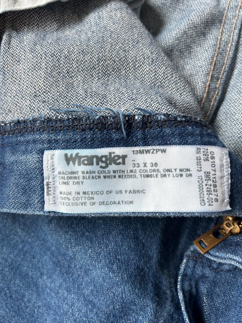 May include: Close-up of a pair of blue denim Wrangler jeans. The label reads "Wrangler" with size 33 x 36. The tag also includes washing instructions and fabric details. A brass zipper is visible.
