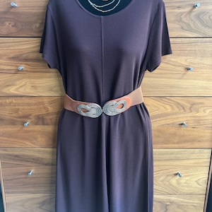 May include: A dark brown dress with short sleeves and a brown belt with a silver buckle. A silver necklace is draped around the neckline. The dress is displayed on a black mannequin against a wooden dresser.