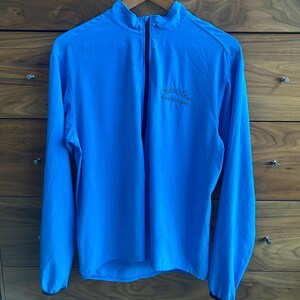 May include: A bright blue athletic jacket with a full-length zipper and a stand-up collar. The jacket has long sleeves and a small logo on the chest. The jacket is hanging on a wooden hanger against a wooden dresser.