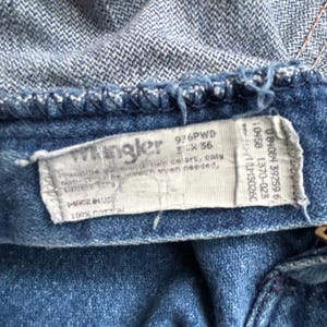 Pu&ograve; includere: Primo piano di jeans blu con una cintura in denim blu chiaro. &Egrave; visibile un'etichetta bianca con il marchio "Wrangler" e altro testo. I jeans sono realizzati in 100% cotone.