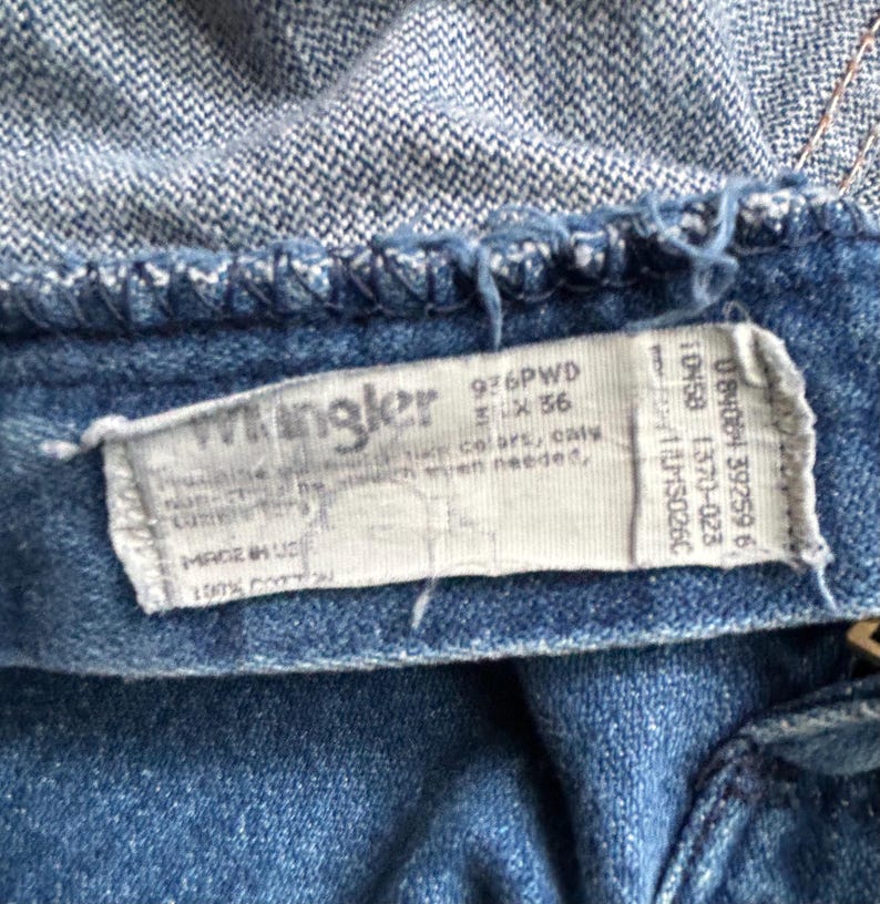 Pu&ograve; includere: Primo piano di jeans blu con una cintura in denim blu chiaro. &Egrave; visibile un'etichetta bianca con il marchio "Wrangler" e altro testo. I jeans sono realizzati in 100% cotone.