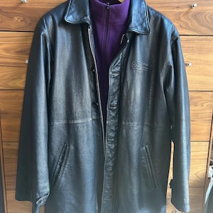 May include: A black leather jacket with a collared neckline and button-down closure. The jacket has two front pockets and a purple fleece jacket underneath. The jacket is hanging on a wooden hanger.