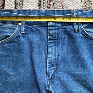 Pu&ograve; includere: Primo piano di jeans blu con un metro a nastro giallo sulla vita. I jeans hanno un classico stile a cinque tasche e una chiusura con bottone e cerniera. La vita misura circa 45.7 cm.