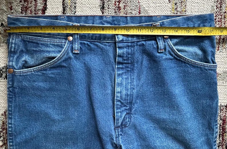 Pu&ograve; includere: Primo piano di jeans blu con un metro a nastro giallo sulla vita. I jeans hanno un classico stile a cinque tasche e una chiusura con bottone e cerniera. La vita misura circa 45.7 cm.