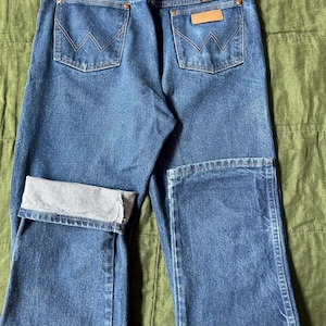 May include: Blue denim jeans with the Wrangler logo on the back pocket. The jeans have a classic five-pocket design, with a folded cuff on one leg. The stitching is a contrasting orange color.