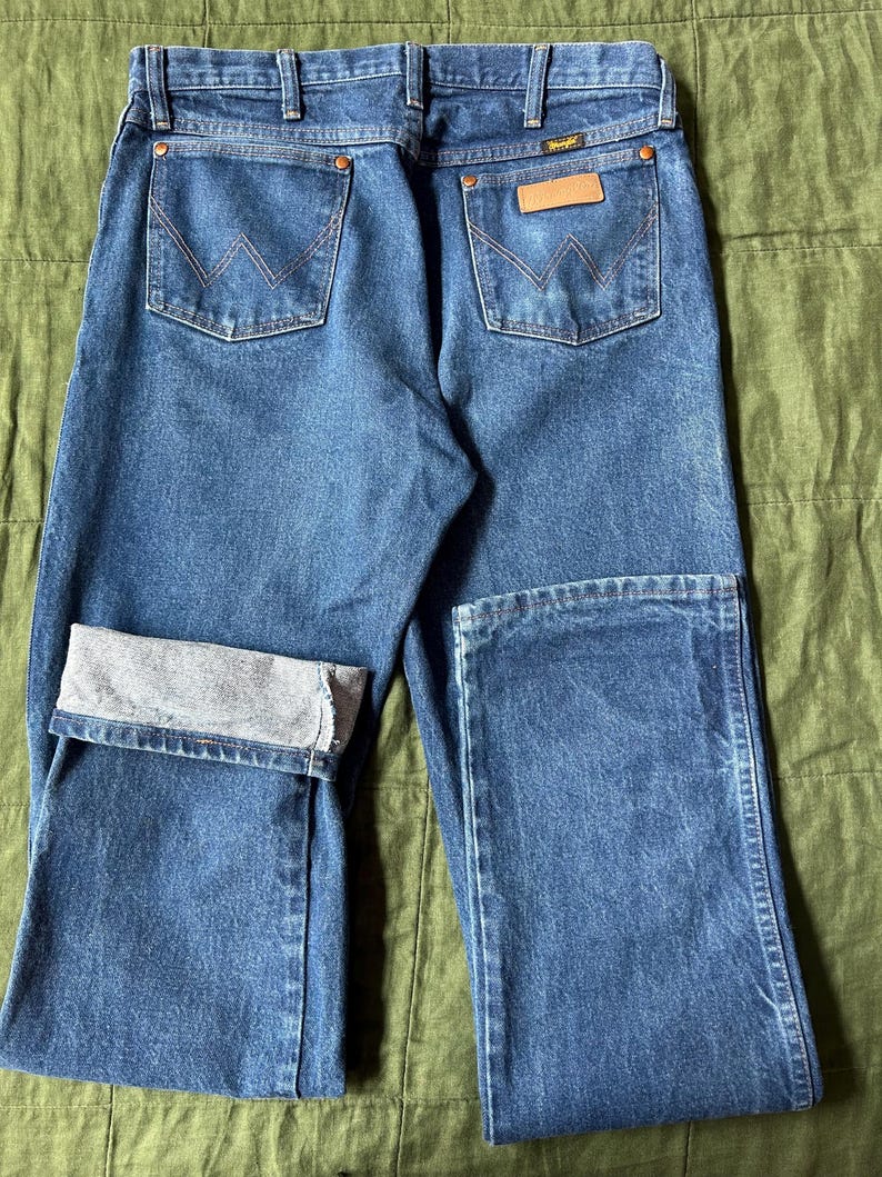 May include: Blue denim jeans with the Wrangler logo on the back pocket. The jeans have a classic five-pocket design, with a folded cuff on one leg. The stitching is a contrasting orange color.