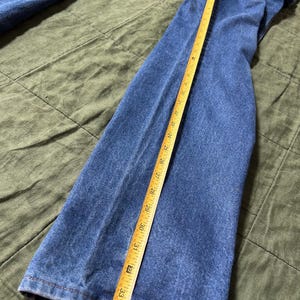 May include: Blue denim jeans with a yellow measuring tape showing a length of approximately 34 inches. The jeans are laid flat on a green surface, showcasing the texture and color of the denim.