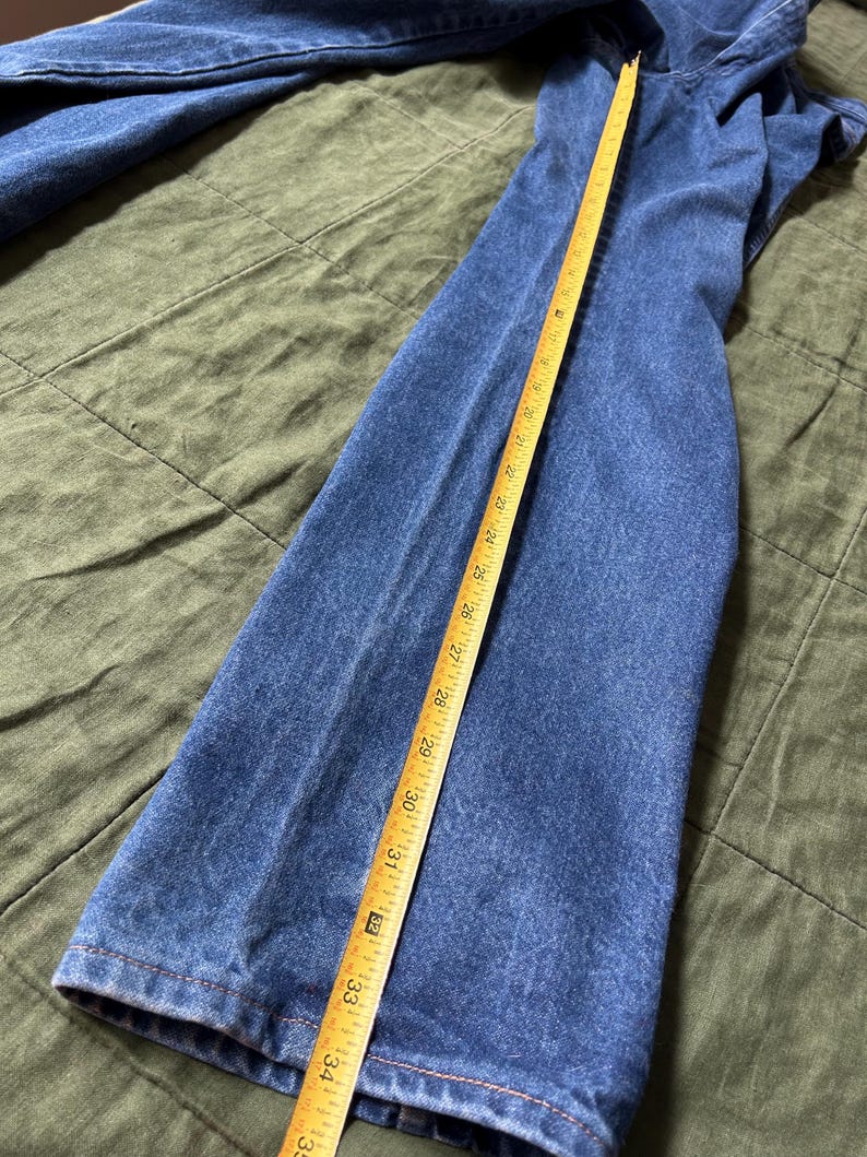 May include: Blue denim jeans with a yellow measuring tape showing a length of approximately 34 inches. The jeans are laid flat on a green surface, showcasing the texture and color of the denim.