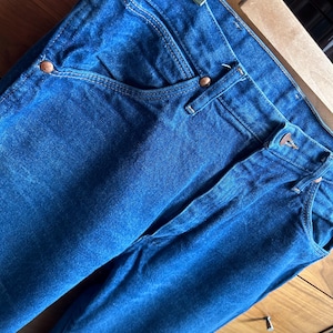 May include: Close-up of blue denim jeans with copper-colored rivets and a classic five-pocket design. The jeans are hanging on a metal and wooden hanger, showcasing the texture and color of the fabric.