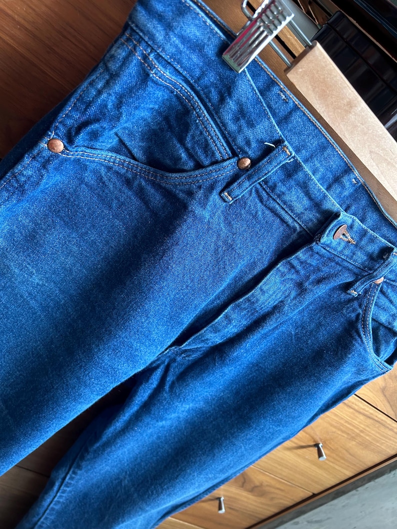 May include: Close-up of blue denim jeans with copper-colored rivets and a classic five-pocket design. The jeans are hanging on a metal and wooden hanger, showcasing the texture and color of the fabric.