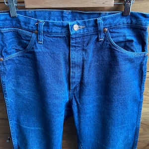 May include: Close-up of blue denim jeans hanging on a metal hanger. The jeans have classic five-pocket styling with copper-colored rivets and a button closure. The denim has a medium wash and a slightly faded appearance.