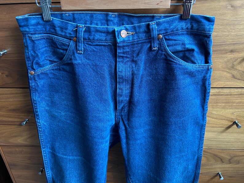 May include: Close-up of blue denim jeans hanging on a metal hanger. The jeans have classic five-pocket styling with copper-colored rivets and a button closure. The denim has a medium wash and a slightly faded appearance.