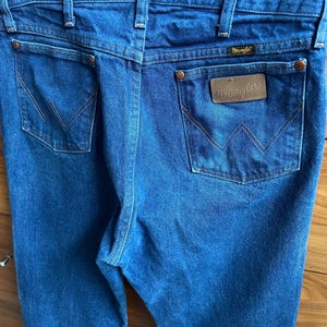 May include: Close-up of blue denim jeans with the Wrangler logo on the back pocket. The jeans have classic five-pocket styling, belt loops, and a copper-colored button and rivets. The stitching is a contrasting tan color.