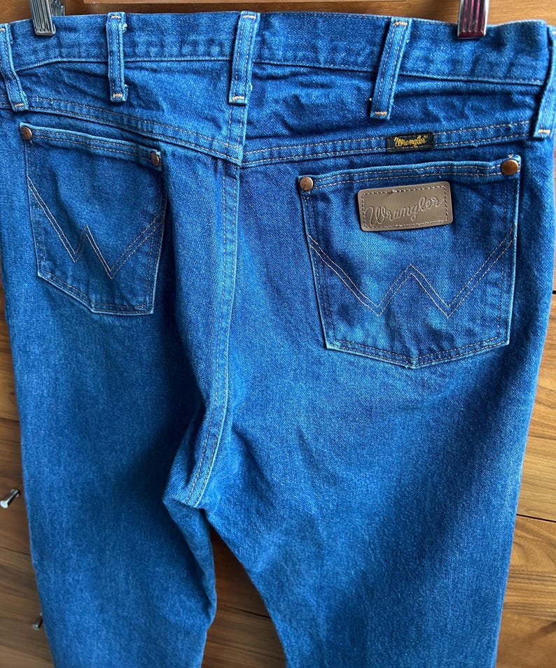 May include: Close-up of blue denim jeans with the Wrangler logo on the back pocket. The jeans have classic five-pocket styling, belt loops, and a copper-colored button and rivets. The stitching is a contrasting tan color.