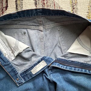 Pu&ograve; includere: Jeans blu in denim con cerniera e chiusura con bottone. Le tasche interne sono visibili, con tessuto bianco e cuciture nere. I jeans hanno un classico design a cinque tasche e un'etichetta Wrangler.