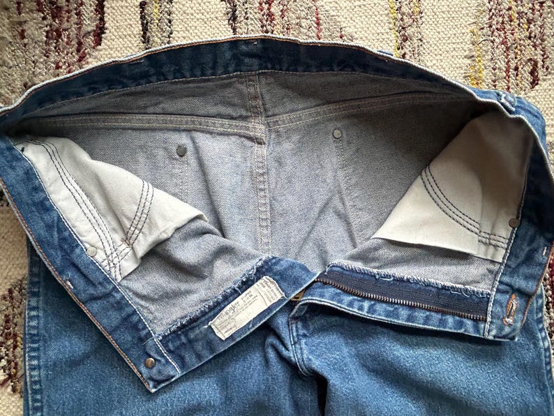 Pu&ograve; includere: Jeans blu in denim con cerniera e chiusura con bottone. Le tasche interne sono visibili, con tessuto bianco e cuciture nere. I jeans hanno un classico design a cinque tasche e un'etichetta Wrangler.