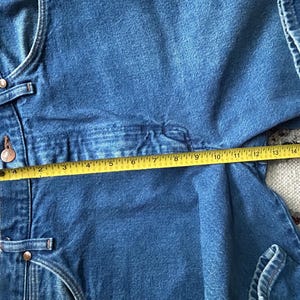 Pu&ograve; includere: Primo piano di una tuta in denim blu con un metro a nastro giallo che mostra una misura di circa 40 cm. La tuta ha bottoni e rivetti in metallo. Il tessuto denim &egrave; di un lavaggio medio.
