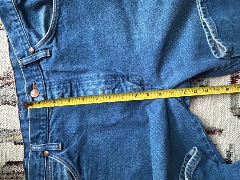 Pu&ograve; includere: Primo piano di una tuta in denim blu con un metro a nastro giallo che mostra una misura di circa 40 cm. La tuta ha bottoni e rivetti in metallo. Il tessuto denim &egrave; di un lavaggio medio.