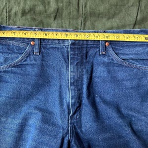 May include: Close-up of blue denim jeans with a yellow measuring tape across the waist. The tape shows a measurement of approximately 16 inches. The jeans have classic five-pocket styling with copper rivets.