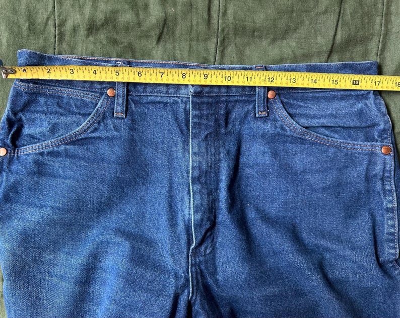 May include: Close-up of blue denim jeans with a yellow measuring tape across the waist. The tape shows a measurement of approximately 16 inches. The jeans have classic five-pocket styling with copper rivets.