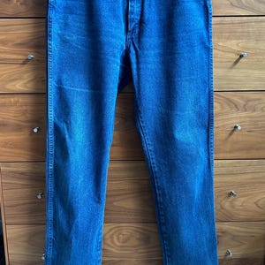 May include: Blue denim jeans with a classic five-pocket design. The jeans are a medium wash with a straight leg cut. The jeans are hanging on a wooden hanger.