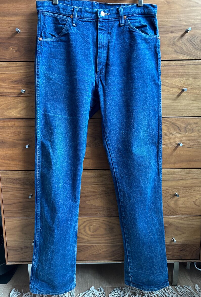 May include: Blue denim jeans with a classic five-pocket design. The jeans are a medium wash with a straight leg cut. The jeans are hanging on a wooden hanger.