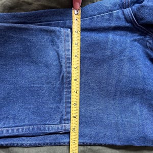 May include: Close-up of blue denim fabric being measured with a yellow tape measure. The tape measure shows a length of approximately 13 inches. The denim is on a green surface.
