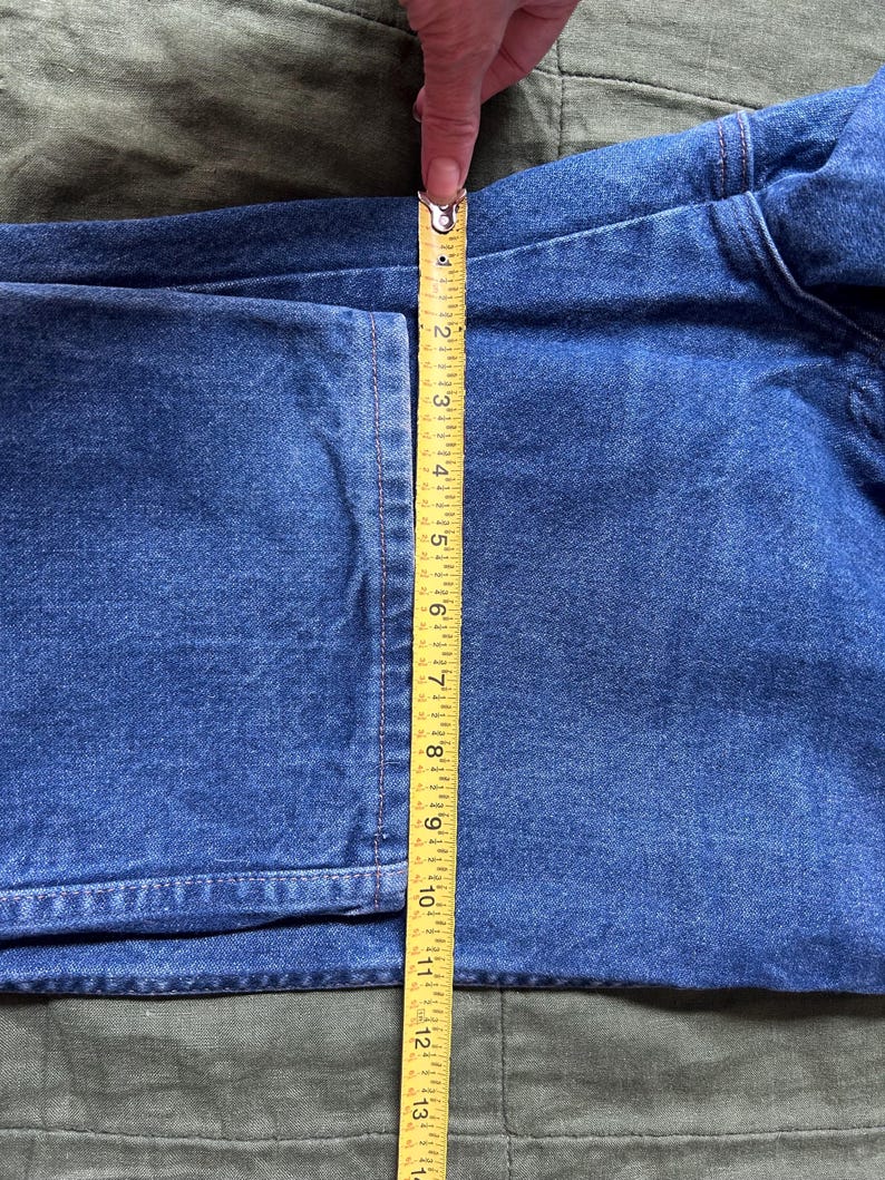 May include: Close-up of blue denim fabric being measured with a yellow tape measure. The tape measure shows a length of approximately 13 inches. The denim is on a green surface.