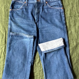 May include: Blue denim jeans with a classic five-pocket design. The jeans have a button and zipper closure, with copper-colored rivets and a folded cuff on one leg. The denim has a faded appearance.