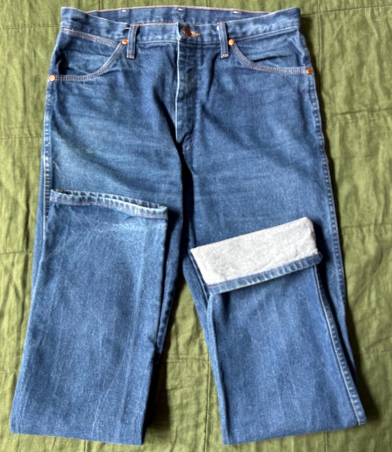 May include: Blue denim jeans with a classic five-pocket design. The jeans have a button and zipper closure, with copper-colored rivets and a folded cuff on one leg. The denim has a faded appearance.