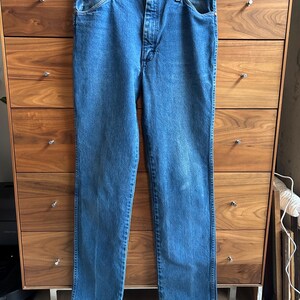 Pu&ograve; includere: Jeans blu in denim appesi a una gruccia di legno. I jeans hanno un classico design a cinque tasche, una chiusura con bottone e cerniera e una vestibilit&agrave; dritta. Il denim ha un lavaggio medio con qualche sbiadimento.