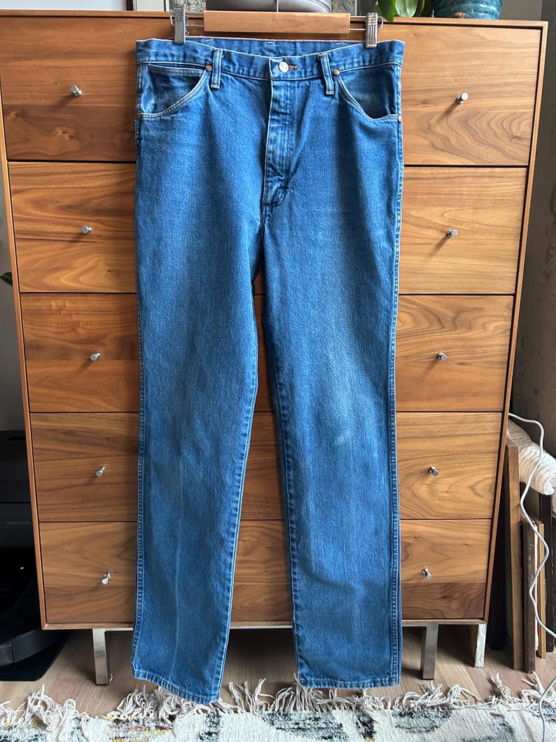 Pu&ograve; includere: Jeans blu in denim appesi a una gruccia di legno. I jeans hanno un classico design a cinque tasche, una chiusura con bottone e cerniera e una vestibilit&agrave; dritta. Il denim ha un lavaggio medio con qualche sbiadimento.