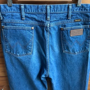 Pu&ograve; includere: Primo piano del retro di jeans Wrangler in denim blu. I jeans hanno due tasche posteriori con il logo Wrangler su un'etichetta in pelle marrone. I jeans sono appesi a una gruccia di metallo.