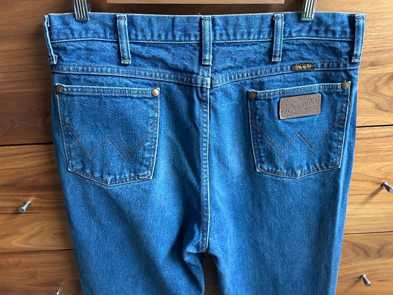 Pu&ograve; includere: Primo piano del retro di jeans Wrangler in denim blu. I jeans hanno due tasche posteriori con il logo Wrangler su un'etichetta in pelle marrone. I jeans sono appesi a una gruccia di metallo.