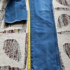 Pu&ograve; includere: Jeans blu in denim con design a gamba dritta. Un metro a nastro giallo &egrave; posto accanto ai jeans, mostrando una lunghezza di circa 86 cm. I jeans sono appoggiati su un tappeto fantasia.