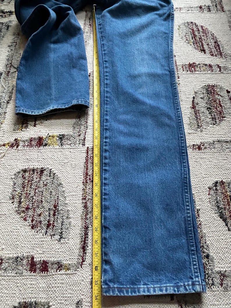 Pu&ograve; includere: Jeans blu in denim con design a gamba dritta. Un metro a nastro giallo &egrave; posto accanto ai jeans, mostrando una lunghezza di circa 86 cm. I jeans sono appoggiati su un tappeto fantasia.