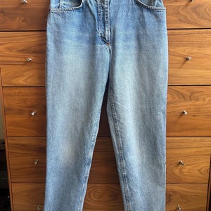 May include: High-waisted light blue denim jeans featuring a classic five-pocket design, a metal button closure, and a zipper fly. The denim has a faded wash with subtle whiskering at the hips and straight-cut legs with reinforced stitching.