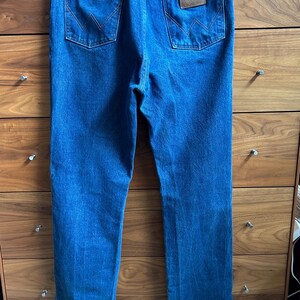 May include: Blue denim jeans with two back pockets and a brown leather patch with the Wrangler logo. The jeans are hanging on a wooden dresser. The jeans have a classic, straight-leg fit.