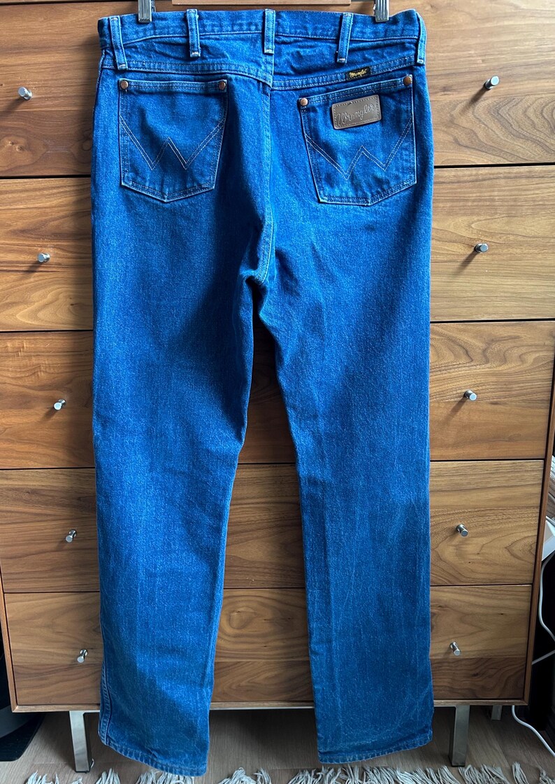 May include: Blue denim jeans with two back pockets and a brown leather patch with the Wrangler logo. The jeans are hanging on a wooden dresser. The jeans have a classic, straight-leg fit.